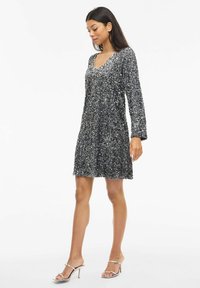 Īsa melna kleita ar spīdumiem, garām piedurknēm, V veida kaklu un izplatītu apakšu, kombinēta ar metāliskām, lencītēm apautām augstpapēžu kurpēm.