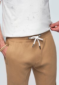 Béžové bavlněné tepláky s elastickým pasem, stahovací šňůrkou a postranními kapsami. V kombinaci s bílým tričkem s jemnými šedými skvrnami.