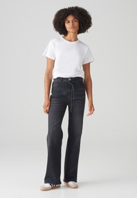 Jean noir taille haute avec une coupe décontractée, présentant un détail de taille nouée, associé à un t-shirt blanc basique et des chaussures décontractées.