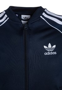 Tummansininen vetoketjullinen takki, joka on valmistettu kankaan kanssa, jossa on valkoisia raitoja. Ominaisuuksiin kuuluu korkea kaulus ja brodeerattu Adidas-logo rinnassa.