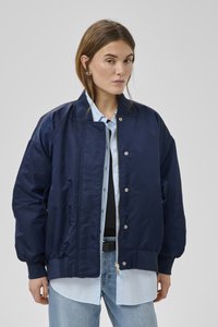 Frau trägt übergroße marineblaue Bomberjacke über hellblauem Hemd und hellblauer Jeans vor einfarbigem grauem Hintergrund.