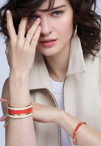 Bracelets en or et corail avec des accents blancs ; un large avec des bandes épaisses et un étroit. Gilet en beige doux avec une fermeture éclair.