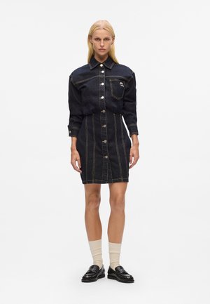 Vestido de camisa de denim en azul oscuro con costuras naranjas que contrastan, frente con botones, cuello y bolsillo en el pecho. Combinado con zapatos y calcetas negras.