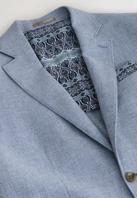 Svetlomodré sako s textúrovaným vzorom tkania, ktoré má vzorovanú tmavomodrú podšívku a zapínanie na jeden gombík. Obsahuje ozdobné vrecko na hrudi.