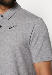 Graues Kurzarm-Polohemd aus strukturiertem Stoff, mit einem schwarzen Nike-Logo auf der linken Brust und einem Knopfkragen.