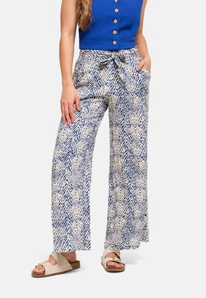 Mujer con pantalones de tiro alto y pierna ancha con estampado de zigzag azul y beige, cinturón atado, top azul sin mangas y sandalias beige.