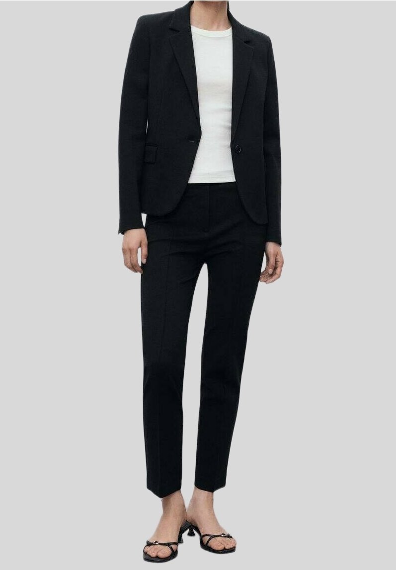 Personne portant un blazer noir ajusté, un pantalon noir slim, une chemise blanche et des sandales noires à bout ouvert, debout devant un fond gris uni.