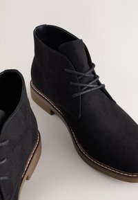 Bottines en daim noir avec un bout arrondi, des lacets fins et un détail de semelle cousue. Caractérisées par une texture lisse et un design minimaliste.