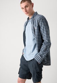 Tenue décontractée avec une chemise en flanelle à carreaux bleus à manches longues sur un polo bleu clair et un short cargo noir. Tissu en coton ; coupe décontractée ; poches.