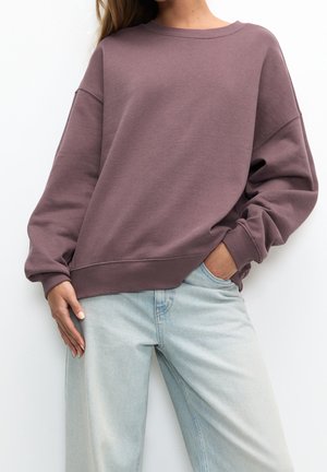Mujer con sudadera oversized color malva con puños y bajo acanalados, combinada con vaqueros de mezclilla de talle alto en azul claro, mano en el bolsillo.