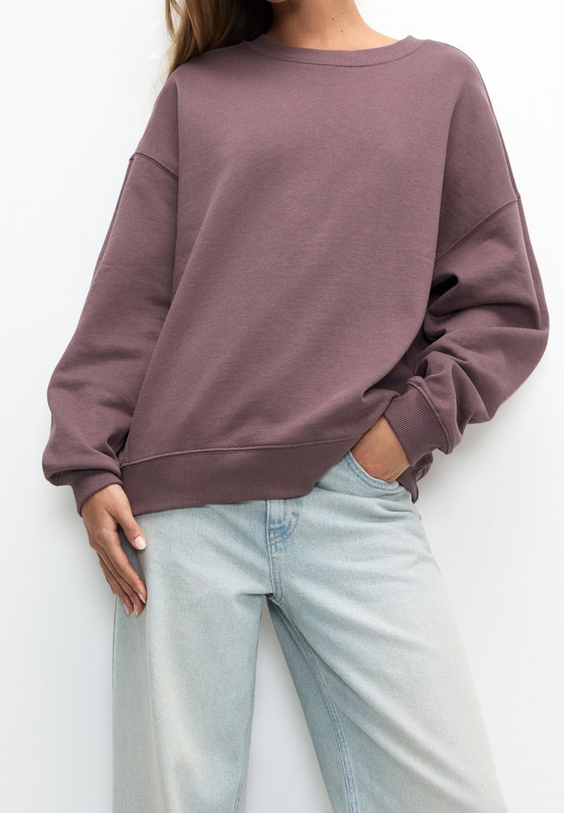 Femme portant un sweat-shirt mauve oversize avec poignets et ourlet côtelés, associé à un jean en denim bleu clair taille haute, main dans la poche.