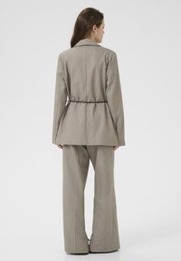 Woman with long brown hair wearing beige tailored suit with flared pants and black belt, standing facing away on plain white background.
