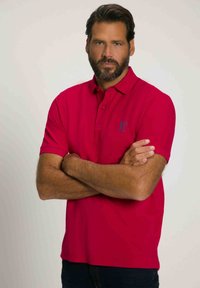 Chemise polo rouge avec un col classique et deux boutons, arborant un logo bleu discret sur la poitrine gauche ; manches courtes et texture lisse.