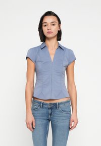 Μπλούζα με κοντά μανίκια σε μπλε χρώμα, εφαρμοστή, με γιακά, φερμουάρ στο μπροστινό μέρος και λεπτομέρειες με πιέτες, συνδυασμένη με ανοιχτόχρωμο τζιν παντελόνι.