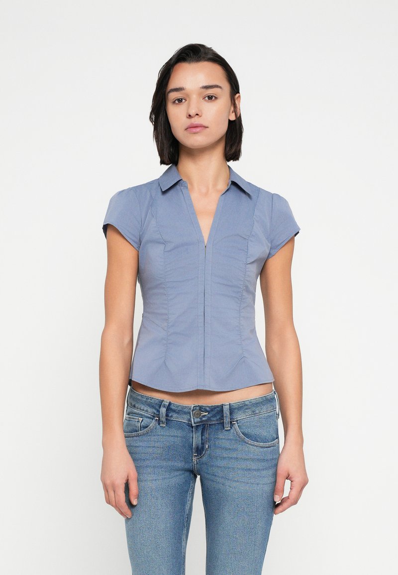 Μπλούζα με κοντά μανίκια σε μπλε χρώμα, εφαρμοστή, με γιακά, φερμουάρ στο μπροστινό μέρος και λεπτομέρειες με πιέτες, συνδυασμένη με ανοιχτόχρωμο τζιν παντελόνι.