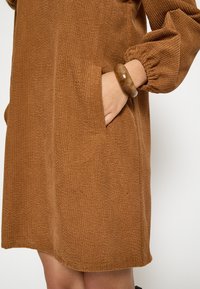 Hnedé manžetové šaty s textúrovaným povrchom, s bočnou kapsou a voľnými elastickými manžetami, doplnené o hrubý náramok.