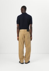 Polo shirt blu scuro con maniche corte abbinato a pantaloni in velluto a coste beige larghi con un'etichetta nera sul retro.