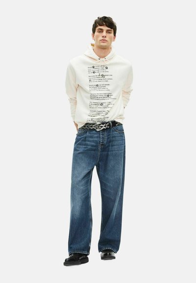 Kapuzensweatshirt in Creme mit bedrucktem Text, kombiniert mit lockeren blauen Jeans und einem schwarzen gemusterten Gürtel, ergänzt durch schwarze Schuhe.