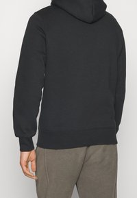 Persona in felpa con cappuccio nera e pantaloni grigi, mostrata di spalle con il braccio sinistro rilassato lungo il fianco.