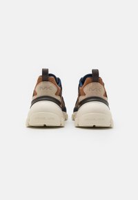 Zapatillas de ante marrón y beige con tela azul marino, suela blanca gruesa, lengüetas de tirón y logotipo "MK" en el talón, vista trasera sobre fondo blanco.