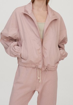 Personne portant une veste zippée rose clair avec des poignets élastiques et des cordons, associée à un pantalon rose clair assorti et un haut beige en dessous.