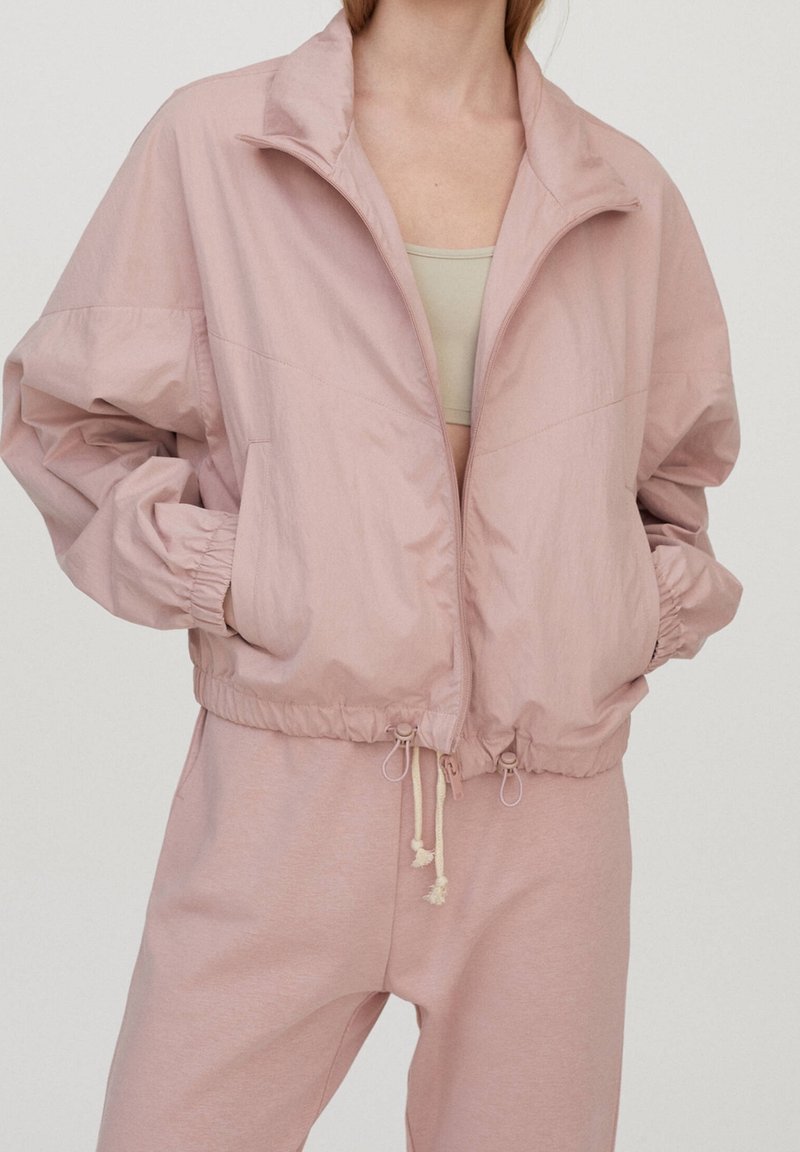 Personne portant une veste zippée rose clair avec des poignets élastiques et des cordons, associée à un pantalon rose clair assorti et un haut beige en dessous.
