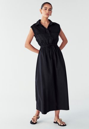 Juodas be rankovių suknelė su mygtukų priekiu, dviem krūtinės kišenėmis, suspaustu liemeniu ir laisva siluetu. Derinama su juodais sandalais, turinčiais auksines detales.