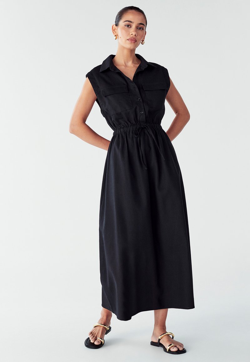 Juodas be rankovių suknelė su mygtukų priekiu, dviem krūtinės kišenėmis, suspaustu liemeniu ir laisva siluetu. Derinama su juodais sandalais, turinčiais auksines detales.