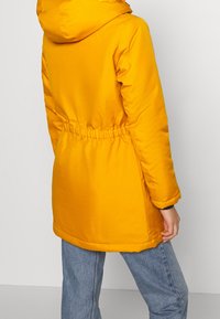 Personne portant un manteau d'hiver à capuche jaune vif avec taille cintrée, associé à un jean bleu, se tenant devant un fond clair uni.