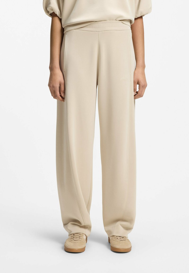 Pantalon beige à jambes larges associé à des baskets beige assorties, porté avec un haut ample beige clair sur un fond blanc.