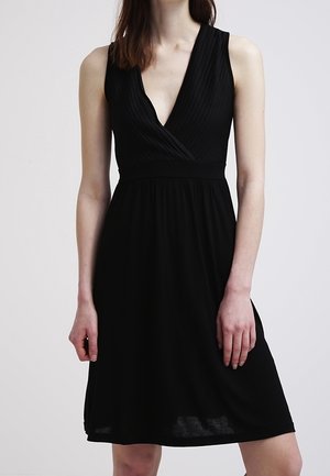 Femme portant une robe noire sans manches arrivant aux genoux, avec un décolleté en V et un corsage plissé, debout devant un fond clair uni.