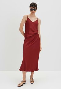 Rotes Satin-Kleid mit dünnen Trägern und einem fließenden Rock. Verfügt über ein dezentes drapiertes Design und eine glatte Textur. Getragen mit flachen Sandalen und Sonnenbrille.