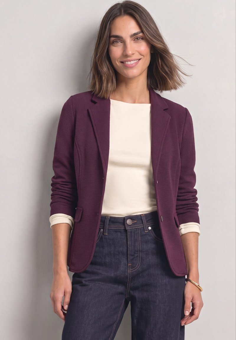 Femme aux cheveux bruns mi-longs portant un blazer violet, un haut crème et un jean bleu foncé, debout devant un fond gris clair.