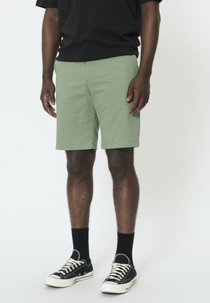 Shorts vert clair à coupe droite, en mélange de coton, avec poches latérales. Associés à des baskets noires et des chaussettes.
