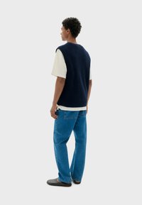 Un gilet en tricot navy sur un t-shirt crème, associé à un jean en denim bleu. Modèle de dos, montrant une coupe décontractée et un style casual.