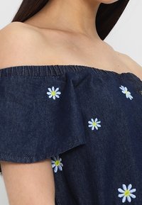 Dark denim off-the-shoulder top with elastic neckline, featuring white daisy embroidery and green accents scattered across the fabric.