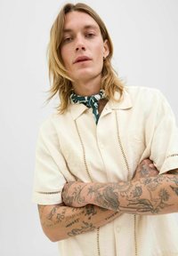 Camisa beige de manga corta con detalles recortados y botón en la parte delantera. El modelo lleva una bufanda verde con estampado y tiene los brazos tatuados cruzados.