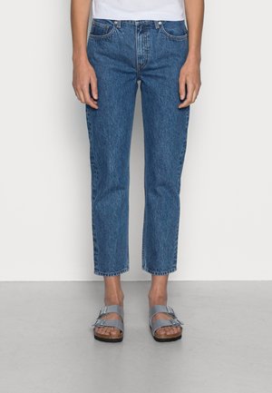 Osoba ubrana w krótkie niebieskie dżinsy i srebrne sandały z podwójną klamrą stoi przy gładkiej białej ścianie i szarej podłodze.