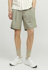 Shorts vert olive clair en matériau doux, avec une coupe droite, des poches latérales et un ourlet net. Associés à des baskets blanches et des chaussettes beiges.