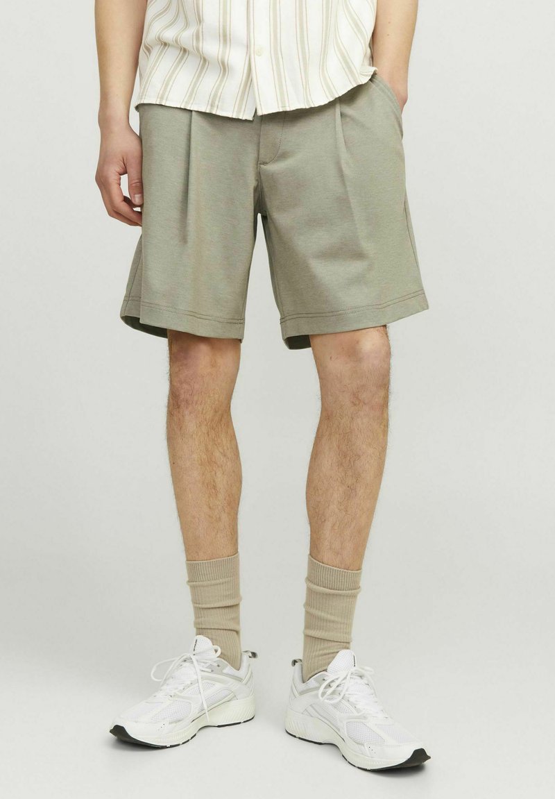 Shorts vert olive clair en matériau doux, avec une coupe droite, des poches latérales et un ourlet net. Associés à des baskets blanches et des chaussettes beiges.