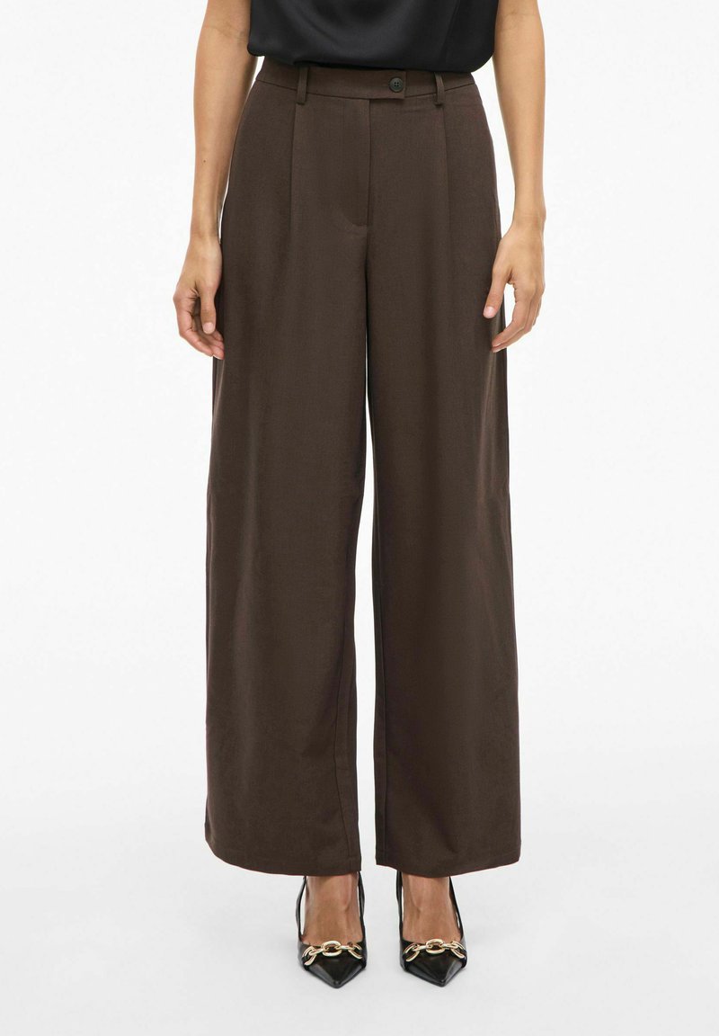 Žena stojaca pred jednoduchým pozadím, oblečená vo vysoký pás široké tmavohnedé nohavice a čierne lodičky špicatého tvaru so zlatým reťazovým detailom.