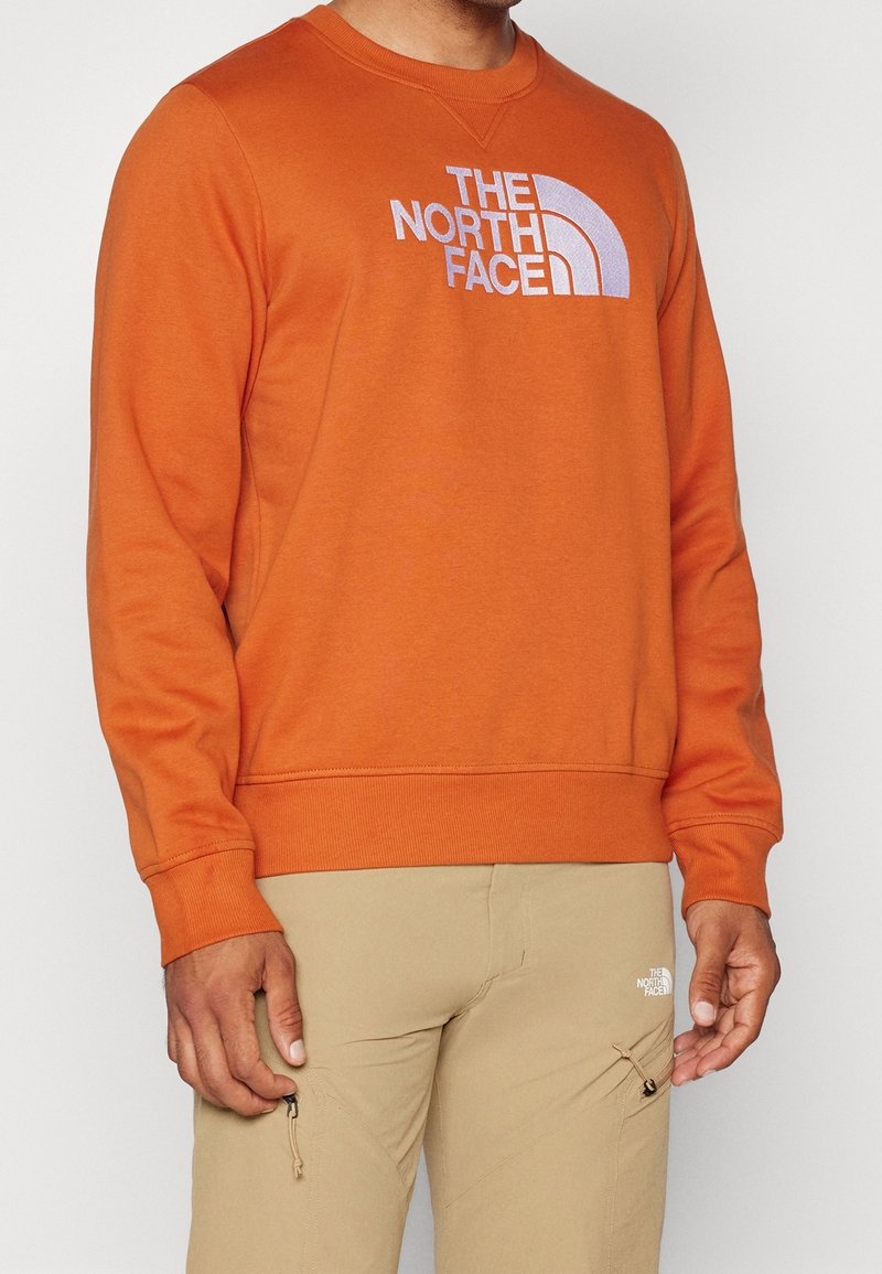 Homme portant un sweat-shirt orange The North Face et un pantalon beige avec le logo The North Face sur la cuisse, debout devant un fond uni.