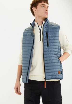 Lyseblå, quilted vest med høj krave, frontlynlås og sidelommer. Båret over en cremet sweater med et logo patch nederst til venstre.