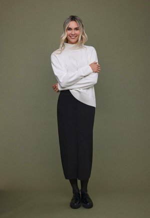 Weißer gestrickter Rollkragensweater mit strukturierten Rautenmustern, kombiniert mit einem schwarzen Midirock und schwarzen Plateauschuhen.