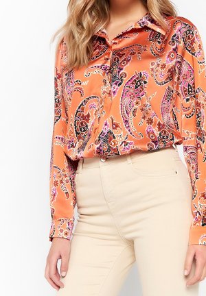 Kvinde iført en langærmet orange paisley mønstret satinbluse, der er stoppet ned i beige bukser med høj talje, vist fra skuldre til hofter.