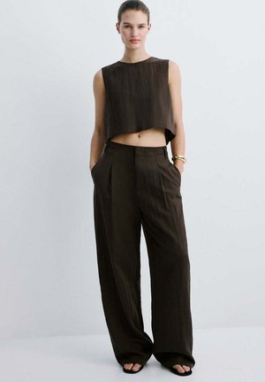 Femme debout, les mains dans les poches, portant un top court sans manches et un pantalon large taille haute en tissu marron foncé sur fond clair.