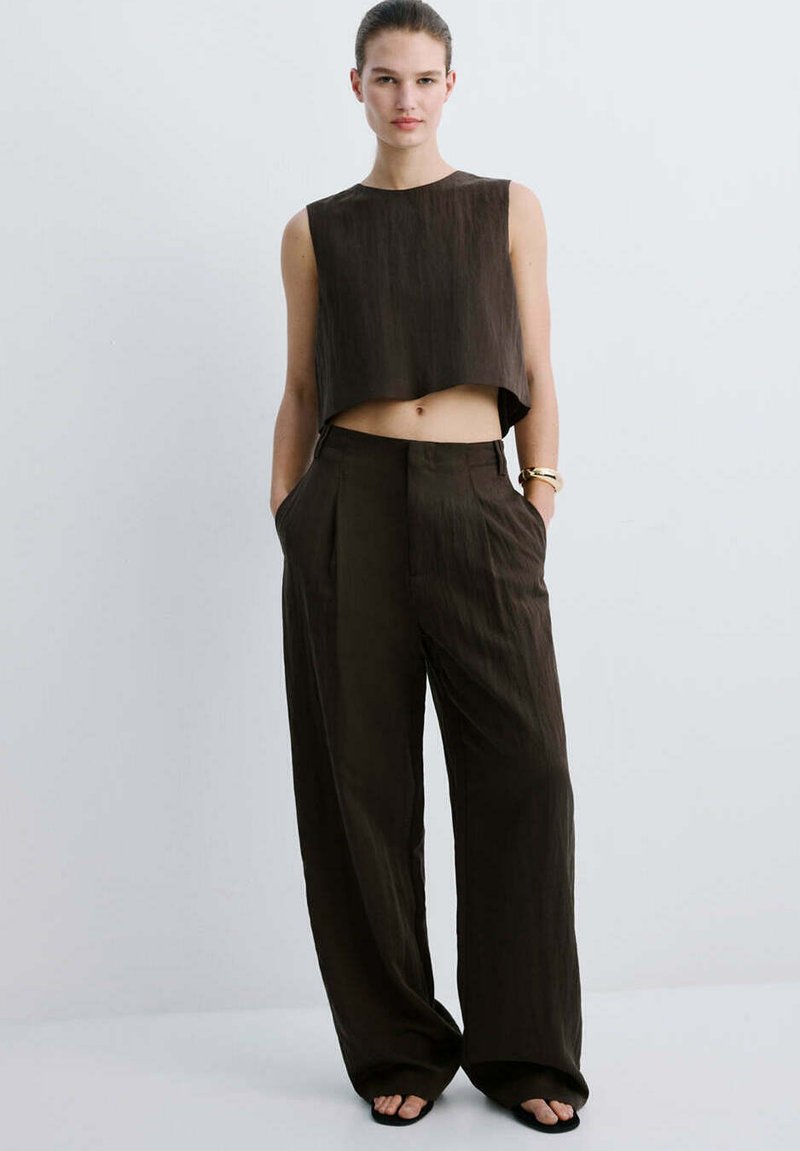 Femme debout, les mains dans les poches, portant un top court sans manches et un pantalon large taille haute en tissu marron foncé sur fond clair.