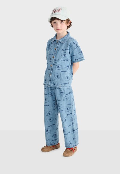 Jeune garçon portant une chemise et un pantalon en denim bleu clair avec des imprimés "Bobo Choses", des chaussures beiges avec des lacets rouges, et une casquette blanche avec du texte rouge.