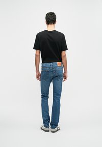 Homem vestindo uma t-shirt preta, jeans azul e tênis brancos está de costas contra um fundo claro e liso.
