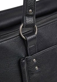 Dettaglio di una borsa in pelle nera con superficie strutturata, accessori metallici, un attacco ad anello e rivetti sulla tracolla per una maggiore durabilità.